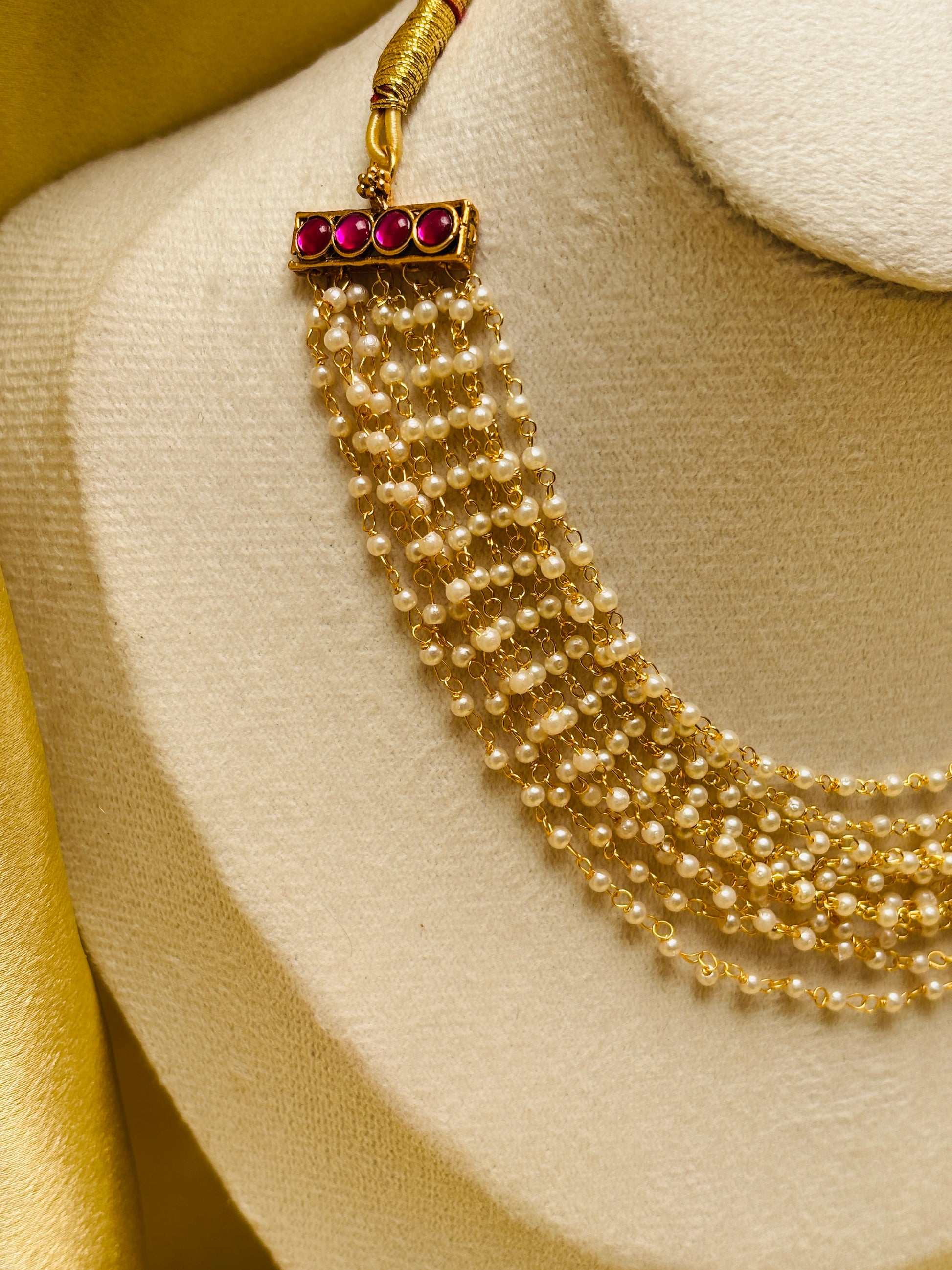 A gold-toned multi-strand pearl necklace with a rectangular gold clasp adorned with four pink gemstones, displayed on a cream-colored jewelry bust.