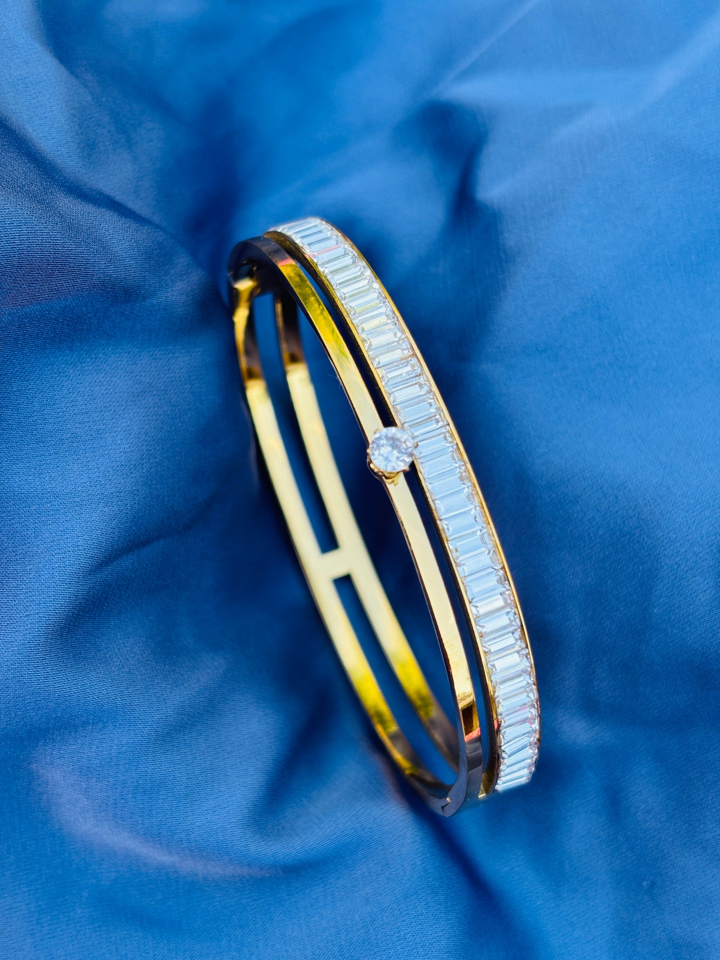 A double-band gold bangle bracelet with a row of baguette-cut clear stones on one side and a single round clear stone at the center, displayed on a textured blue fabric background.